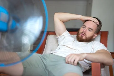 Young bearded man using electric fan at home, sitting on couch cooling off Stock Photos