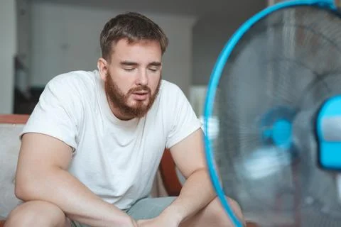 Young bearded man using electric fan at home, sitting on couch cooling off Stock Photos