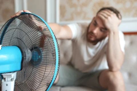 Young bearded man using electric fan at home, sitting on couch cooling off Foto stock