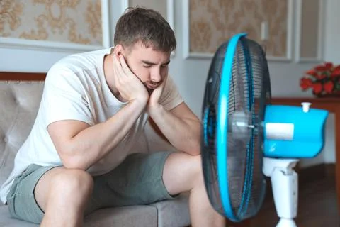 Young bearded man using electric fan at home, sitting on couch cooling off Stock Photos