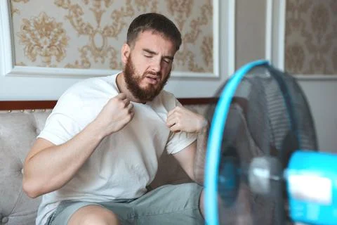 Young bearded man using electric fan at home, sitting on couch cooling off Stock Photos