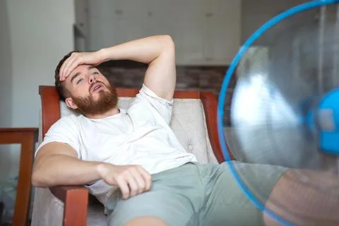 Young bearded man using electric fan at home, sitting on couch cooling off Stock Photos