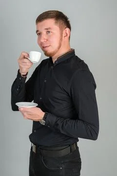 A young bearded red-haired man drinks coffee from a small white mug standing  Stock Photos