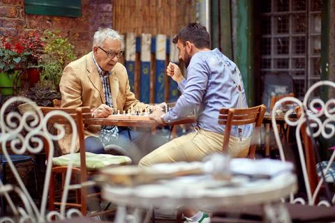 Young beardy guy playing chess with elderly man, sitting in outdoor cafe Stock Photos