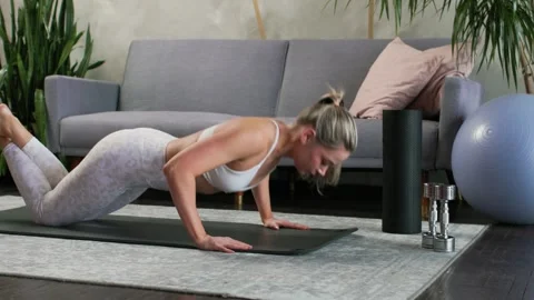 Young beautiful blonde doing knee push-ups on yoga mat in cozy living room. Gray Vídeos de archivo 304403070