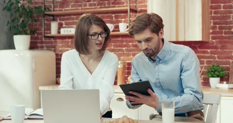 Young beautiful business lady with laptop and husband with tablet, sitting in Video stock 166323115