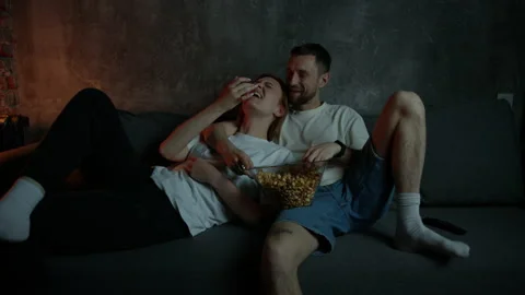 A young beautiful couple is lying on the couch, eating popcorn and watching TV Stock Footage 220563412