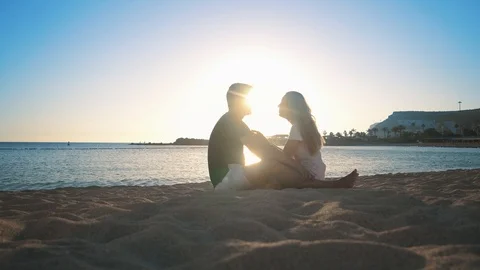 Young beautiful couple is sitting on the sandy shore near the sea, laughing and Stock Footage 104059440