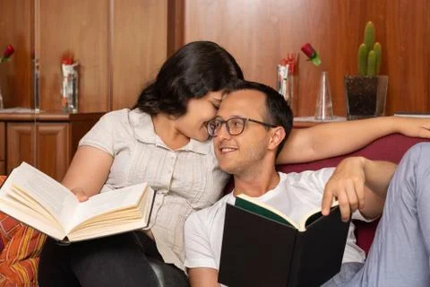 Young beautiful couple whispering while reading a book on the sofa at home Stock Photos