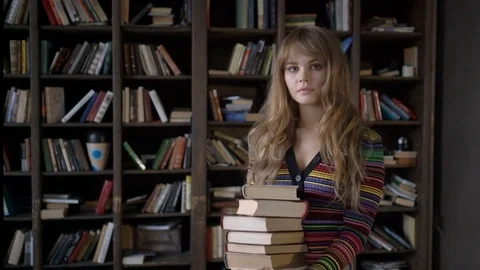 Young beautiful girl holding a stack of books in front of a bookcase Stock Footage 77752438