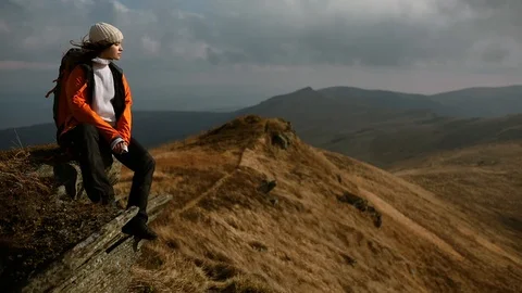 A young beautiful girl in an orange jacket with a backpack sits on a rock and Stock Footage 119585553