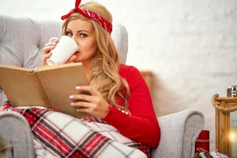 Young beautiful woman reading a book in an armchair with blanket and tea duri Stock Photos