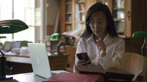 Young biologist is using smartphone sitting at desk with laptop in library. Stock Footage 93581370