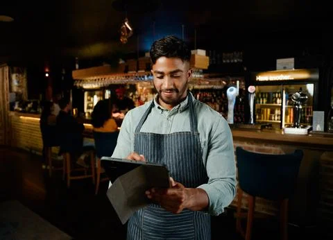 Young biracial man in apron using digital tablet while working as waiter in bar Stock Photos