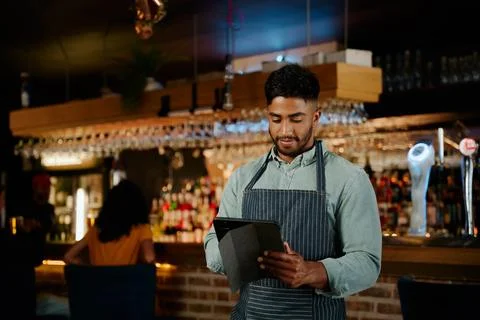 Young biracial man in apron using digital tablet while working as waiter in Stock Photos