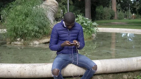Young black african guy making a call wearing earphones in the park 스톡 동영상 104973437