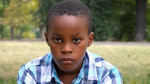 A young  black boy sits on grass in a park and looks seriously at the camera - 스톡 동영상 98290519