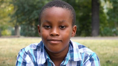 A young  black boy sits on grass in a park and smiles at the camera - face Video stock 98290548