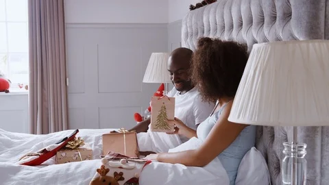 Young black couple sitting up in bed giving gifts to each other and embracing on Vídeo Stock 99763867