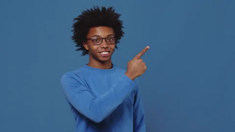 Young black guy in glasses standing on a blue background in the studio, pointing Stock Footage 255941904