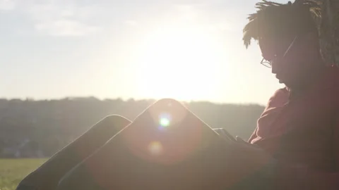 Young black male using a digital tablet as he sits by a tree, in slow motion 스톡 동영상 153027568