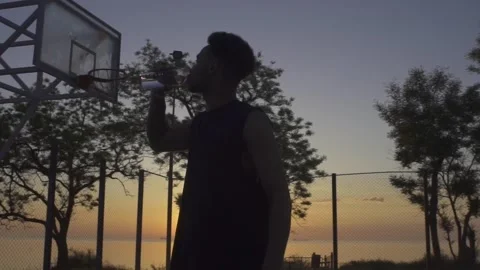 Young black man drinking water while playing basketball on playground on sunrise Stock Footage 160476776