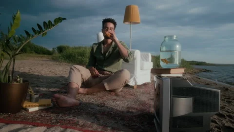 Young Black Man Eating Burger in Living Room Interior on Beach at Sunset Stock Footage 304847258