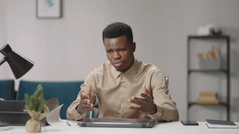 Young black man is sitting at table in room, talking and gesticulating, looking Stock Footage 150662893
