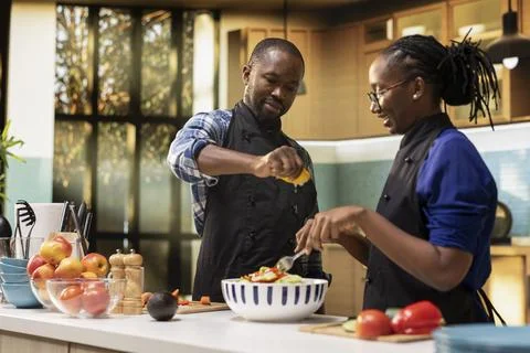 Young black man squeezing lemon juice over veggies and woman mixing ingredients Fotos Stock