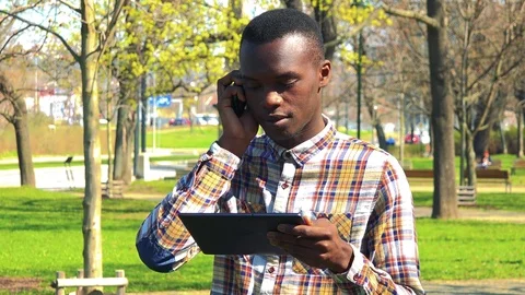 A young black man talks on a smartphone while looking at a tablet in a park on a Stock-Footage 78429115