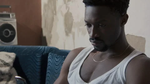 Young Black Man Texting on Flip Phone while Sitting on Sofa in Worn Apartment Видео 329573616