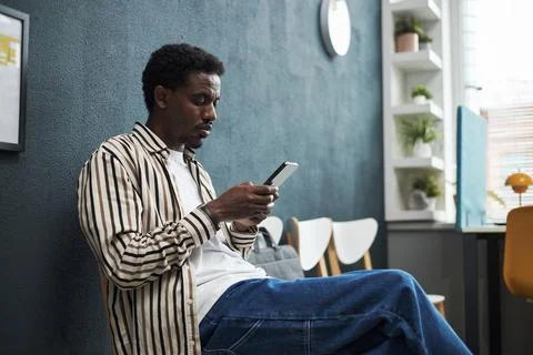 Young Black Man Using Smartphone While Sitting in Queue in Bank Office Stock Photos
