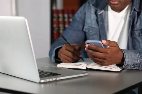 Young black man writing notes in library, using smartphone and computer Foto stock