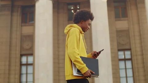 Young black programmer walking outdoors with a laptop and copybook and scrolling Stock Footage 262614105