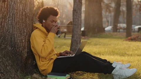 Young black programmer in a yellow hoodie sitting under a tree outdoors and Stock Footage 262072869
