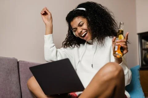 Young black woman in headphones drinking bear while using laptop Stock Photos