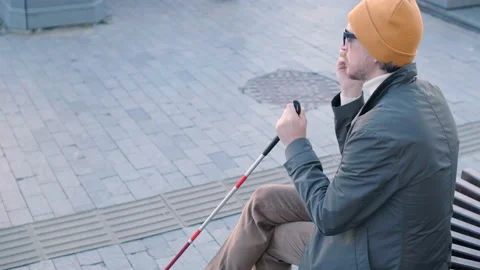 Young blind man with smartphone sitting on bench in park in city Stock Footage 220839238
