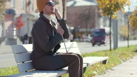Young blind man with smartphone sitting on bench in park in city Stock Footage 221366191