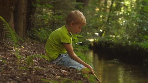 Young Blond Boy Sits by the Brook and Plays with the Wooden Stick. Vídeo Stock 115996270