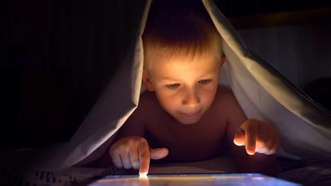 Young blond boy under covers playing on tablet a computer game. Stock Footage 91232637