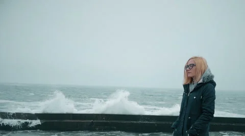 Young blond girl by the beach while cold and rainy watching on the waves Stock Footage 63263643
