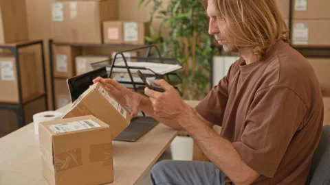 Young blond man scans parcel label with smartphone, hand on box and focused.. Stockbeeldmateriaal 339194734