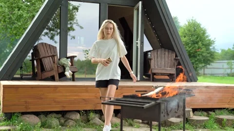 Young Blonde Girl Sitting On The Terrace, Goes To Barbecue Throwing Firewood Stock Footage