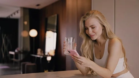 A young blonde in the kitchen drinks water from a glass and chats by phone Stockbeeldmateriaal 161387415
