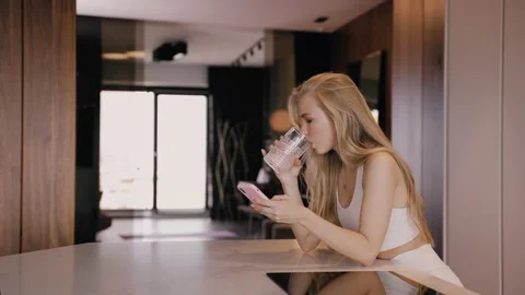 A young blonde in the kitchen smiling, drinks water and chats by the phone Stock Footage 161387639