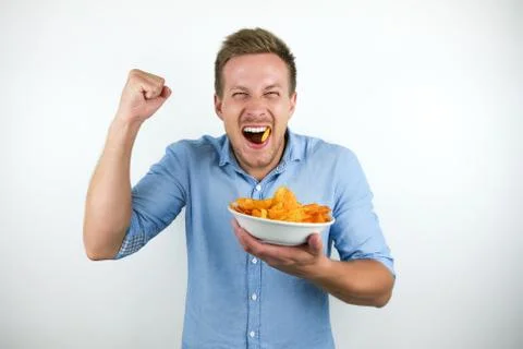 Young blonde man eats chips holding his fist up happily on isolated white Stock Photos