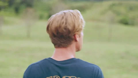 Young blonde man walks through a field, away from camera, towards trees on an ov Stock Footage 199461205