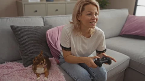 Young blonde woman playing video games on a couch with her chihuahua dog in.. Vídeos de archivo 278270953