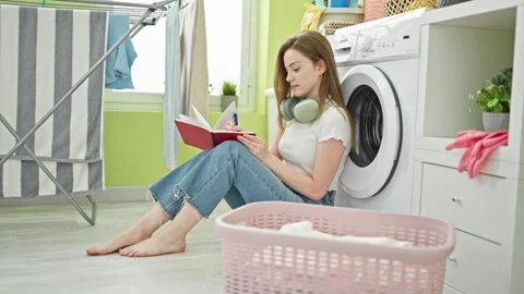 Young blonde woman taking notes waiting for washing machine at laundry room Stock Footage 247377940