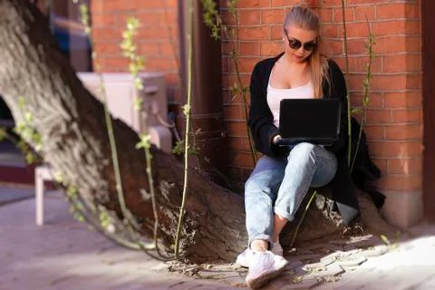 A young blonde working at a computer while sitting on a tree near a brick wal Stock Photos